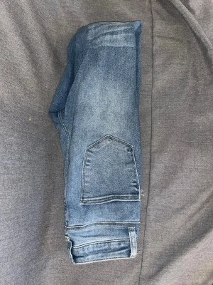 Blå slitna jeans från Akira - Säljer ett par blå jeans från Akira med snygga slitningar och klassisk femficksdesign. Jeansen har en rak passform och är tillverkade i denim med en lätt tvättad look. Perfekta för en avslappnad och trendig stil.