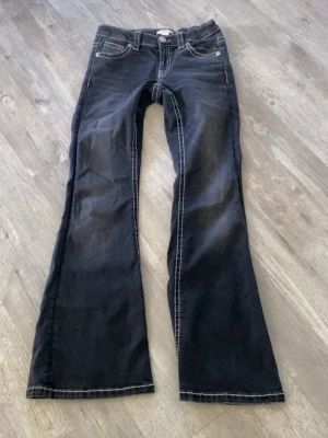 Mörkgråa bootcut jeans med kontrastsömmar - Snygga mörkgråa jeans med bootcut passform och vita kontrastsömmar. Jeansen har klassiska fem fickor och dekorativa sömmar på bakfickorna. Perfekta för dig som gillar en retroinspirerad look.