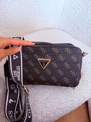 Svart axelväska från Guess - Snygg svart axelväska från Guess med guldiga logomönster och metalltriangel framtill. Väskan har bred, justerbar axelrem med texten 'Guess Los Angeles' i vitt. Två dragkedjefack och detaljer i guld. Tillverkad i konstläder.