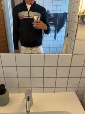 Svart vindjacka från BOSS - Snygg svart vindjacka från BOSS med dragkedja framtill och rutigt mönster över bröstet. Jackan har vita och röda detaljer samt hög krage. Perfekt för dig som vill ha en stilren och sportig look.