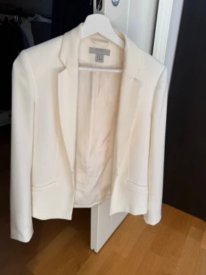 Vit kavaj från H&M, storlek 34 - Stilren vit kavaj från H&M i storlek 34. Kavajen har klassisk passform med två fickor framtill och är helfodrad. Materialet är strukturerat och ger en snygg look. Perfekt för dig som vill ha en enkel och elegant kavaj.