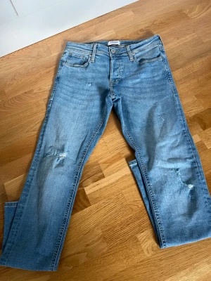 Ljusblå raka jeans med slitningar - Säljer ett par ljusblå jeans med raka ben och snygga slitningar på både lår och knä. Klassisk femficksmodell med knapp och dragkedja framtill. Perfekta till en avslappnad stil och passar till det mesta. 30/32 slim Glenn 