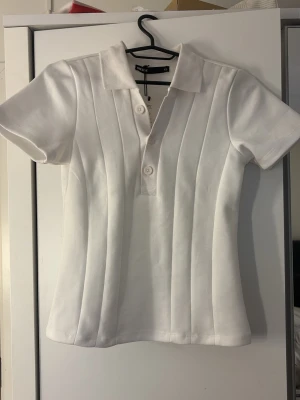 Bikbok topp - Klassisk vit polotopp från bikbok med ribbade vertikala ränder och korta ärmar. Toppen har krage och knappar framtill för en sportig look. Perfekt till jeans eller kjol för en clean och fräsch stil.