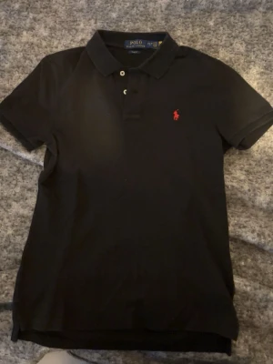 Svart pikétröja från Ralph Lauren - Klassisk svart pikétröja från Ralph Lauren med röd broderad logga på bröstet. Slim fit-modell med korta ärmar, krage och knappar framtill. Perfekt för en stilren och sportig look.