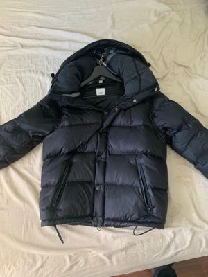 Svart pufferjacka från burberry - Snygg svart pufferjacka från burberry med dragkedja och knappar framtill, två dragkedjeförsedda fickor och justerbar huva med rutigt innerfoder. Jackan har en klassisk boxig passform och är perfekt för kalla dagar. Vill sälja snabbt eftersom ska flytta!