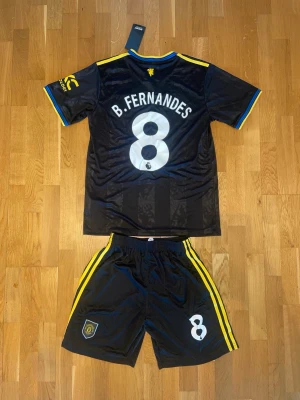 Bruno Fernandes Manchester United bortaställ 23/24 - Snyggt Manchester United bortaställ från säsongen 23/24 med B. Fernandes och nummer 8 på ryggen. Setet är svart med gula detaljer, Adidas-logga och Snapdragon sponsortryck. Tröjan har korta ärmar och shorts med gula ränder på sidorna. Perfekt för fotbollsträning eller match.