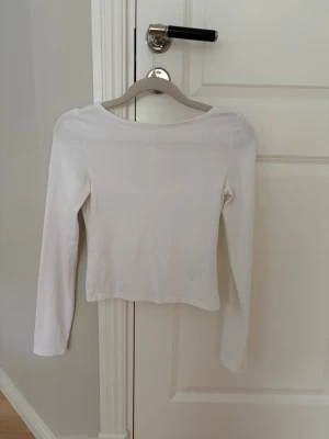 Vit långärmad croppad topp - Säljer en enkel och stilren vit långärmad topp med croppad passform och rundad urringning vid ryggen. Perfekt att matcha med jeans eller kjol för en clean look. Materialet känns mjukt och stretchigt, troligen bomull eller en bomullsblandning.