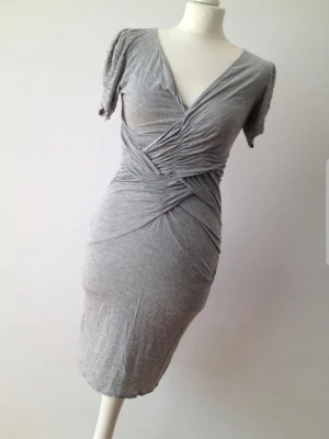 Snygg klänning från All Saints - Supersnygg grå kortklänning från All Saints med omlott och draperad effekt framtill. Puffiga kortärmar med diskreta pärldetaljer ger en unik touch. Mjuk och stretchig bomullskänsla, perfekt för dig som gillar stilrena men ändå speciella plagg.