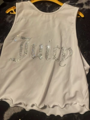 Vit Juicy linnetopp med strass - Säljer en vit linnetopp från Juicy med glittrande strassdetaljer som stavar 'Juicy' på framsidan. Toppen är ärmlös och har vågig kant nertill för en extra snygg touch. Perfekt för dig som gillar statement-plagg!