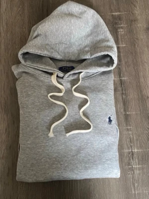 Grå hoodie från Polo Ralph Lauren - Klassisk grå hoodie från Polo Ralph Lauren med vit dragsko i huvan och den ikoniska lilla loggan broderad på bröstet. Hoodien har känguruficka framtill och ribbade muddar vid ärmar och nederkant. Perfekt för en avslappnad och stilren look.