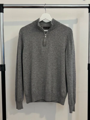 Grå half-zip från Oscar Jacobson - Grå stickad tröja i kashmir från Oscar Jacobson med half-zip. Tröjan har långa ärmar och ribbade ärmslut och nederkant. Den är sparsamt använd och är mycket bra skick