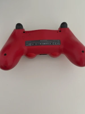 Röd PlayStation 4 DualShock handkontroll - Säljer en röd PlayStation 4 DualShock handkontroll från Sony. Den har klassisk form med två analoga spakar, touchpad och ikoniska PlayStation-knappar. Materialet är plast och färgen är matt röd med svarta detaljer. Perfekt för gaming och multiplayer.
