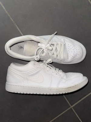 Vita Air Jordan 1 Low sneakers - Säljer ett par helvita Air Jordan 1 Low sneakers från Nike. Skorna har klassisk låg siluett, perforerad tåbox och ikoniska Air Jordan-loggan broderad på hälen. Tillverkade i läder med matchande vita skosnören och sula. Perfekta för en clean och stilren look.