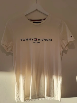 Vit T-shirt från Tommy Hilfiger - Klassisk vit T-shirt från Tommy Hilfiger med tryckt logga och text 'EST. 1985' på bröstet. T-shirten har rund halsringning med randig detalj och korta ärmar. Tillverkad i mjuk bomull för en skön känsla. Liten logga även på ärmen.