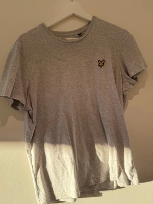 Grå t-shirt från Lyle & Scott - Snygg grå t-shirt från Lyle & Scott med klassisk rund hals och korta ärmar. T-shirten har den ikoniska gula loggan broderad på bröstet och är tillverkad i mjuk bomull för en bekväm känsla. Perfekt till jeans eller shorts.