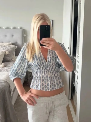 Blommig omlottopp med puffärm - Söt blommig omlottopp med v-ringning och puffiga trekvartsärmar. Toppen har smock i midjan för en figurnära passform och är croppad. Perfekt för dig som gillar romantisk stil och vill ha något luftigt till sommaren.