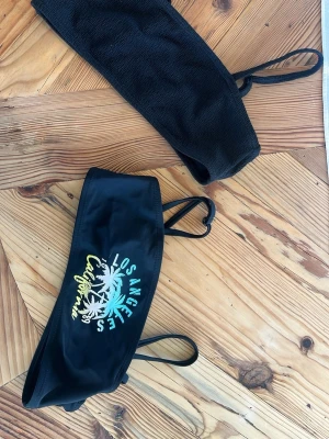 Svart bikini bandeau Los Angeles - Svart bandeau bikini med tryck i turkos, vit och gul text 'Los Angeles California' och palmträd. Ena delen är enfärgad svart med struktur, den andra har tryck och tunna axelband. Perfekt för stranden eller poolen.