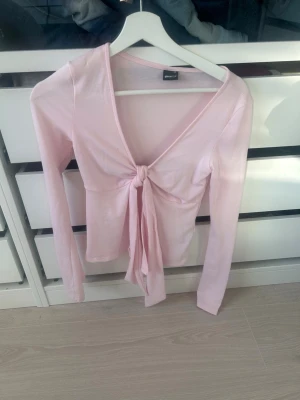 Ljusrosa knytblus från Gina Tricot - Supersöt ljusrosa topp från Gina Tricot med långa ärmar och knytning framtill. Djup V-ringning och mjukt, stretchigt material som sitter snyggt på kroppen. Perfekt för att skapa en trendig och feminin look.