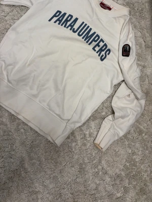 Vit sweatshirt från Parajumpers - Säljer en vit sweatshirt från Parajumpers med stor logga i blått över bröstet och patch på ena ärmen. Tröjan har rund halsringning, ribbade muddar och är tillverkad i mjukt material. Perfekt för en avslappnad och stilren look. ÄKTA!! Vill bara bli av me den