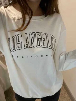 Vit sweatshirt med Los Angeles-tryck - Trendig vit sweatshirt med svart text 'Los Angeles California' på bröstet. Oversized passform och rund halsringning. Perfekt för en avslappnad streetwear-look.