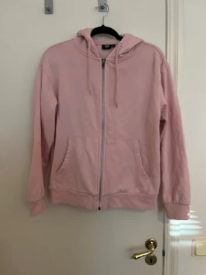 Ljusrosa hoodie med dragkedja - En ljusrosa hoodie med dragkedja framtill, två fickor och justerbar huva med snören. Ribbade muddar vid ärmslut och nederkant. 