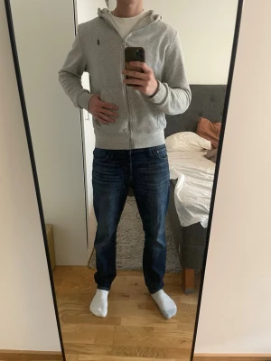 Grå hoodie med dragkedja från Polo Ralph Lauren - Snygg grå hoodie från Polo Ralph Lauren med dragkedja framtill och klassisk huva. Tröjan har två fickor och en liten broderad logga på bröstet. Perfekt för en chill och stilren look. Materialet är mjukt och skönt, passar till det mesta.