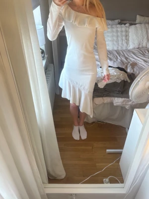 Vit långklänning med volang - Superfin vit långklänning med volangdetalj längs axeln och nederkanten. Klänningen har lång ärm och en elegant, figurnära passform. Perfekt för dig som vill ha en stilren och chic look.