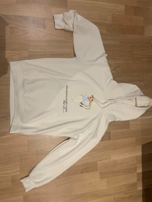 Vit Ralph lauren Hoodie med polo bear tryck  - Säljer en stilren vit hoodie med huva och känguruficka framtill. På bröstet finns ett tryck med en tecknad figur och text. Hoodien har ribbade muddar och är i mjuk material.