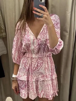 Rosa och vit kortklänning med volang - Söt kortklänning i rosa och vitt med blommigt mönster, v-ringning och volangdetaljer. Klänningen har trekvartsärm och markerad midja med lätt rynk. Perfekt för sommarens varma dagar.