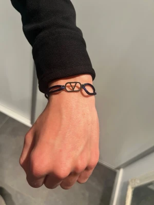 Valentino armband - Snyggt armband från Valentino med svart justerbart snöre och guldfärgat metallspänne i form av det ikoniska V-logotypen. Perfekt accessoar för att lyfta din outfit med en stilren och modern touch.
