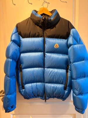 Blå och svart pufferjacka Moncler - Säljer en blå och svart pufferjacka från Moncler med klassisk logga på bröstet. Jackan har hög krage, två dragkedjefickor och quiltad design. Perfekt för kalla vinterdagar och riktigt snygg streetstil.