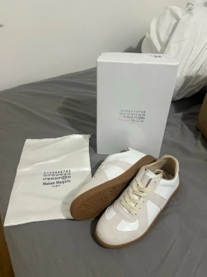 Maison Margiela vita sneakers med mockadetaljer - Maison Margiela sneakers i vitt läder med beige mockadetaljer och klassisk gummisula. Lågt skaft och stilren design med vita snören. Perfekta för dig som gillar minimalistisk och trendig stil.