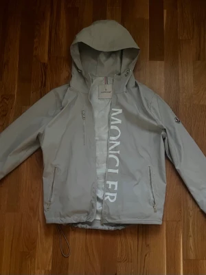 Grå vindjacka från Moncler - Snygg grå vindjacka från Moncler med stor logga längs dragkedjan framtill. Jackan har huva, två dragkedjefickor och justerbar nederkant. Moncler-märke på ärmen och klassiska ränder på insidan av kragen. Perfekt för blåsiga dagar.