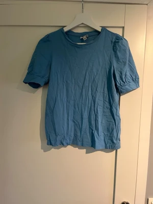 Blå kortärmad topp med puffärm - En stilren blå topp med rund halsringning och korta puffärmar. Tillverkad i mjuk bomull som känns skön mot huden. Perfekt att matcha med jeans eller kjol för en avslappnad look.