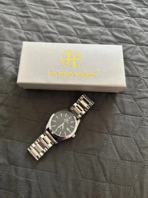 Pablo Raez klocka  - Stilren klocka från Pablo Raez med svart urtavla och silverfärgat armband i metall. Klockan har datumvisning och levereras i originalask med extra länkar och verktyg för justering. Perfekt accessoar för en modern look.
