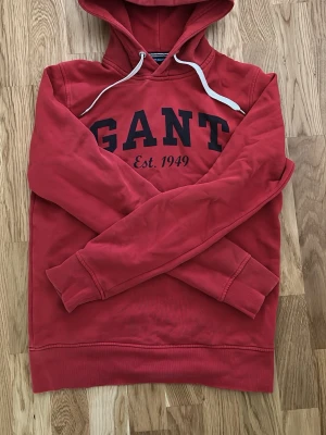 Röd hoodie från GANT med tryck - Röd hoodie från GANT med svart tryck framtill där det står 'GANT Est. 1949'. Klassisk huva med vita snören och ribbade muddar vid ärmar och nederkant. Perfekt för en avslappnad och sportig stil.