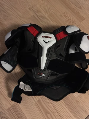 CCM FT6 Pro axelskydd SR Small - Säljer ett par svarta, vita och röda axelskydd från CCM, modell FT6 Pro. Skydden är i seniorstorlek small och har robust konstruktion med extra skydd vid axlar och bröst. Perfekt för hockey eller annan kontaktsport.