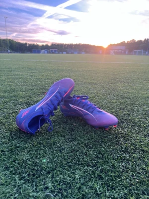 Blå-lila Puma fotbollsskor - Säljer ett par snygga blå-lila fotbollsskor från Puma med röda detaljer och vita loggor på sidorna. Skorna har snörning och är designade för att ge bra grepp på planen. Perfekta för dig som vill sticka ut på matchen med stilrena och sportiga skor.