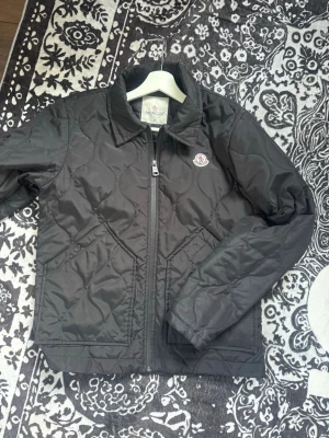 Svart dunjacka från Moncler - Snygg svart dunjacka från Moncler med quiltat mönster och dragkedja framtill. Jackan har två snedställda fickor med tryckknapp och klassisk Moncler-logga på bröstet. Perfekt för kalla vinterdagar.
