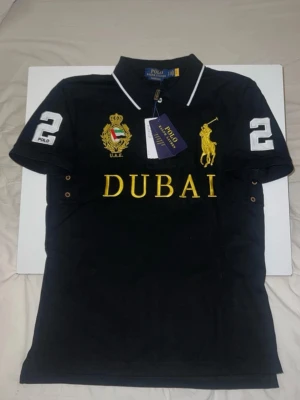  Polo Ralph Lauren - Svart pikétröja från Polo Ralph Lauren med gul broderad logga, texten DUBAI och U.A.E.-emblem på bröstet. Stora vita siffror '2' på ärmar och rygg. Klassisk krage med vit kant och korta ärmar. Perfekt för en sportig och stilren look.
