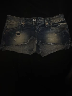 Mörkblå slitna jeansshorts - Säljer ett par mörkblå jeansshorts med slitna detaljer och fransig kant. Shortsen har klassisk femficksdesign, låg midja och råa kanter för en avslappnad look. Perfekta till sommaren för en cool och trendig stil.