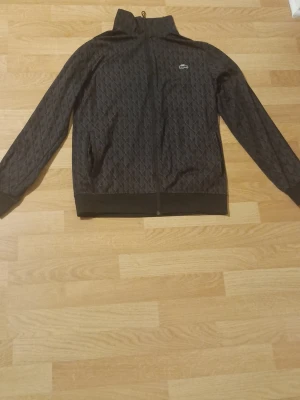 Svart quiltat loungewear-set Lacoste - Snyggt svart loungewear-set från Lacoste med quiltat mönster. Setet består av en zip-jacka med hög krage och matchande byxor med dragsko i midjan. Båda delarna har den klassiska Lacoste-loggan och ribbade muddar. Perfekt för chill dagar hemma.