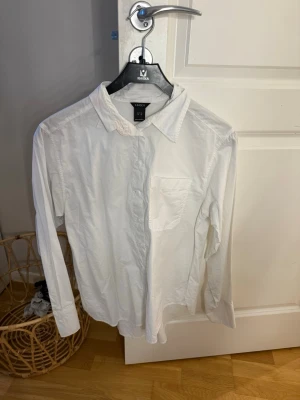 Vit skjorta från Lindex - Klassisk vit skjorta från Lindex med lång ärm och bröstficka. Skjortan har en enkel krage och knäppning framtill. Tillverkad i ett tunt, mjukt material som känns bekvämt mot huden. Perfekt för en clean och stilren look.