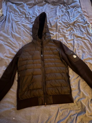 Svart cardigan jacka från Moncler - Snygg svart cardigan jackafrån Moncler med quiltad front och huva. Jackan har släta ärmar, ribbade muddar och dragkedjor i sidorna. Klassisk Moncler-logga på ena ärmen och praktiska fickor. Perfekt för kalla vinterdagar.