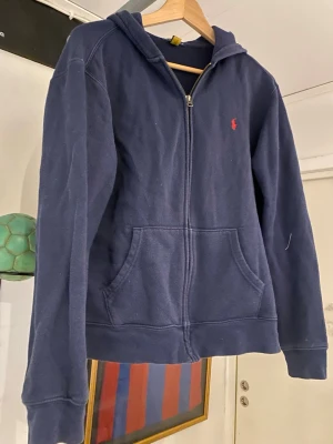 Mörkblå hoodie från Ralph Lauren - Mörkblå hoodie med dragkedja från Ralph Lauren. Klassisk design med huva, kängurufickor och det ikoniska röda Polo-logot på bröstet. Tillverkad i mjukt bomullsmaterial som är skönt att ha på sig. Perfekt för en avslappnad och stilren look.