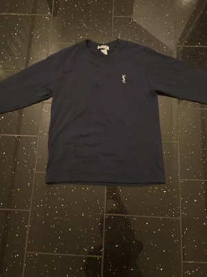 Mörkblå långärmad tröja från YSL - Snygg mörkblå långärmad tröja från Yves Saint Laurent med klassisk vit YSL-logga broderad på bröstet. Tröjan har rund halsringning och är tillverkad i ett mjukt material som känns skönt mot huden.