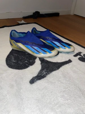 Adidas Messi blå fotbollsskor - Snygga fotbollsskor från Adidas Messi-kollektionen i blått med ljusblå och vita detaljer samt guldiga inslag på hälen. Skorna har en strumpliknande passform och är tillverkade i syntetmaterial för bästa bollkontroll och komfort på planen.