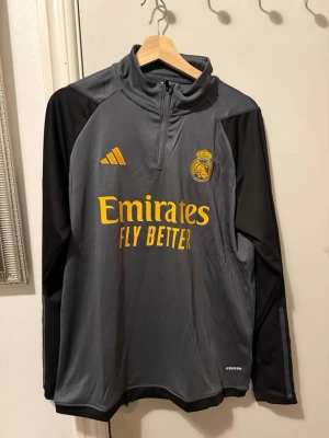 Grå Real Madrid träningströja Adidas - Snygg grå långärmad träningströja från Adidas med Real Madrid-logga och gula detaljer. Tröjan har svart på ärmarna, halv dragkedja och tryckt Emirates Fly Better på bröstet. Perfekt för fotbollsträning eller sportiga dagar.