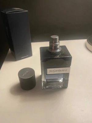 Yves Saint Laurent parfymflaska - Snygg parfym från Yves Saint Laurent i en stilren, fyrkantig glasflaska med svart lock och silverdetaljer. Flaskan har en modern design och etikett med varumärket på framsidan. Perfekt för dig som gillar exklusiva dofter.