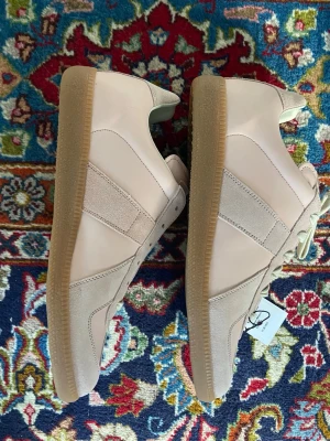 Masion margeila GATs storlek 42  - Snygga beige sneakers med ovandel i både mocka och slätt läder, samt klassisk gummisula. Skorna har ljusa snören och en stilren design som passar till det mesta. Perfekt för dig som gillar minimalistisk och clean stil. Storlek 42 och har mycket mer info, hör av dig!